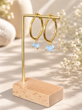 Gold hoop earrings with small light blue bead drops, displayed on a gold stand with a wooden base against a warm neutral background with dried flowers.