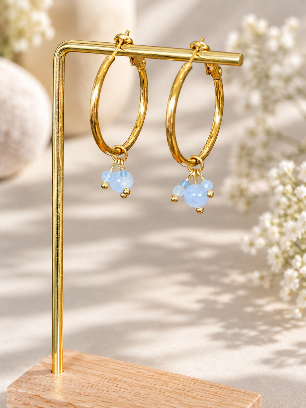 Gold hoop earrings with small light blue bead drops, displayed on a gold stand over a wooden base against a softly blurred beige background with white flowers.