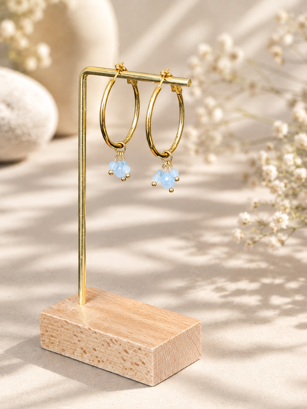 Gold hoop earrings with small light blue bead drops, displayed on a gold stand with a wooden base against a warm beige background with soft floral props.