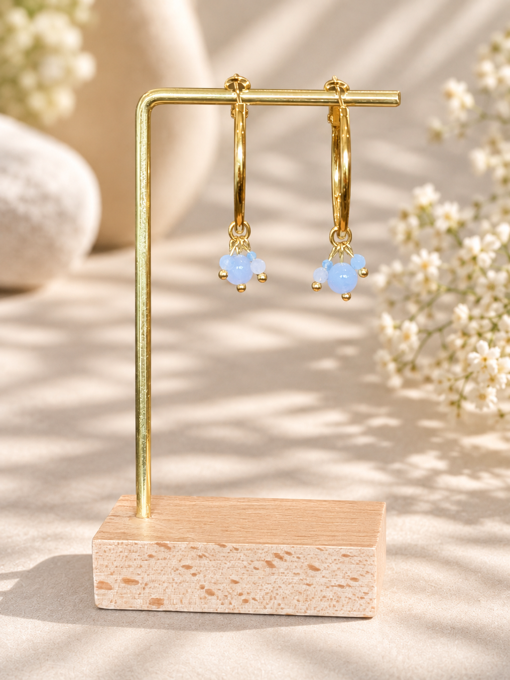A pair of gold hoop earrings with small light blue bead clusters hanging from the hoops, displayed on a gold U-shaped stand with a wooden base against a softly lit neutral background with blurred flowers.