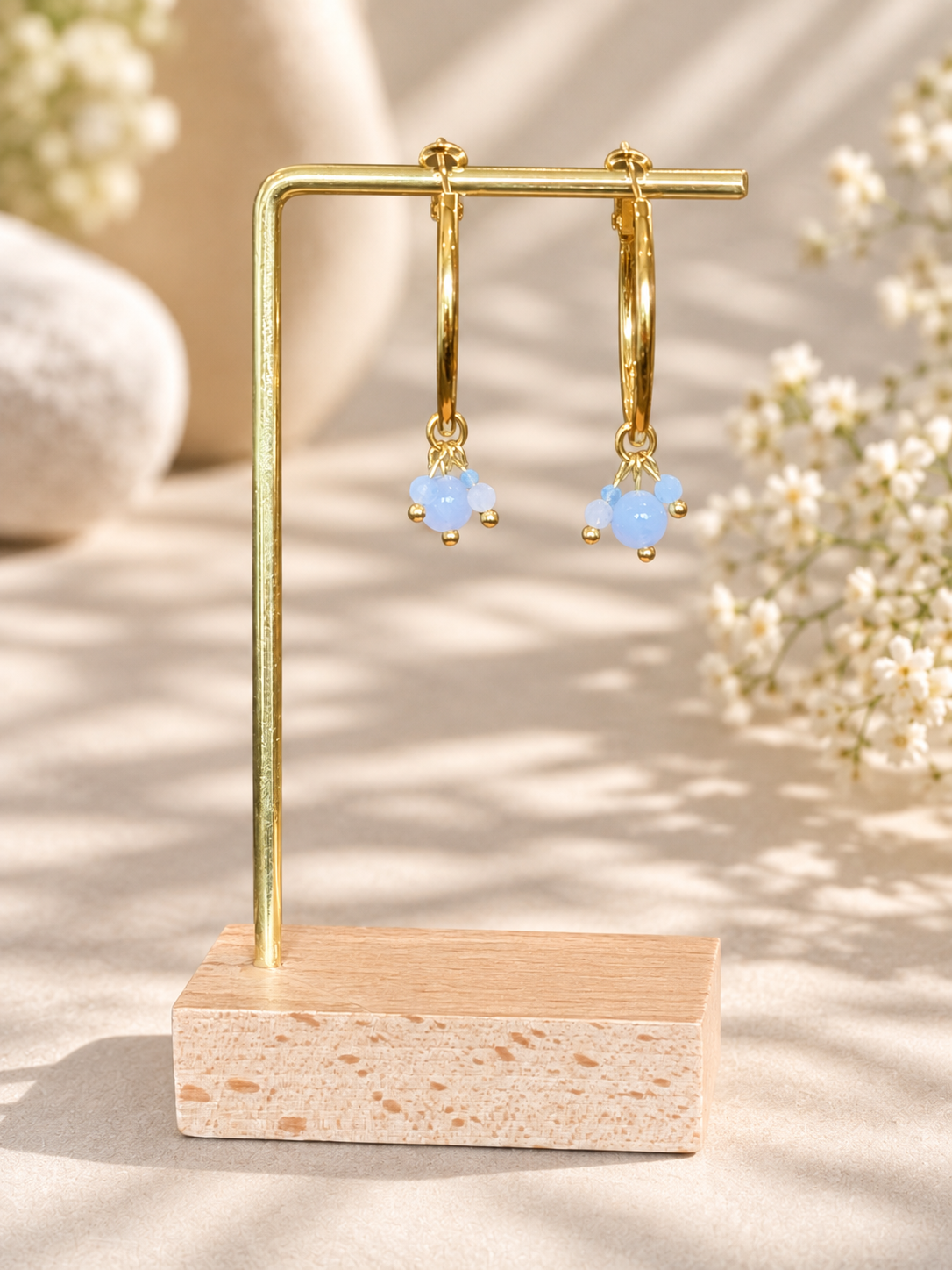 A pair of gold hoop earrings with small light blue bead clusters hanging from the hoops, displayed on a gold U-shaped stand with a wooden base against a softly lit neutral background with blurred flowers.
