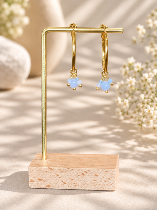 A pair of gold hoop earrings with small light blue bead clusters hanging from the hoops, displayed on a gold U-shaped stand with a wooden base against a softly lit neutral background with blurred flowers.