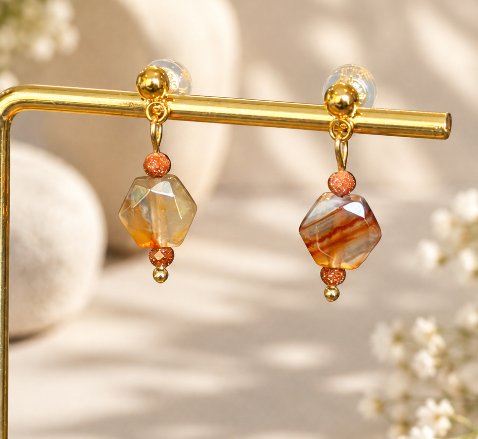 A pair of gold drop earrings with faceted amber and orange beads, displayed on a gold stand against a warm beige background with soft floral bokeh.