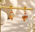 A pair of gold drop earrings with faceted amber and orange beads, displayed on a gold stand against a warm beige background with soft floral bokeh.