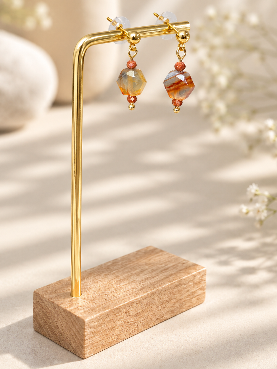 A pair of dangling earrings displayed on a gold-tone stand with a wooden base. The earrings feature faceted amber-orange beads, small copper-toned beads, and clear crystal drops, set against a softly blurred warm neutral background with dried flowers.