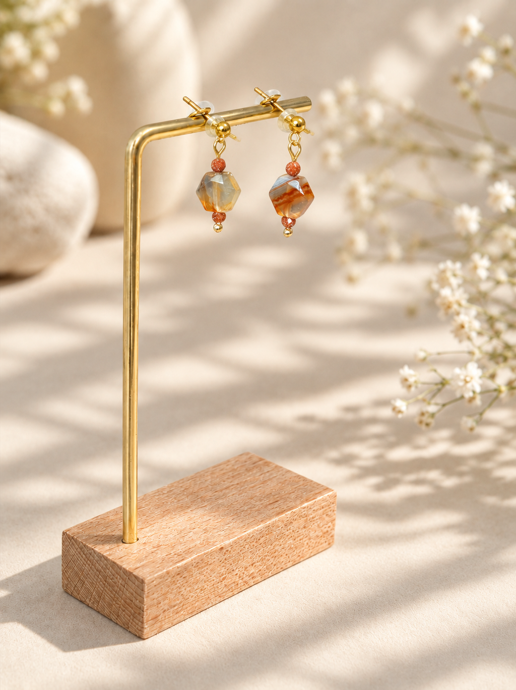 A pair of gold-tone drop earrings with faceted orange-brown beads and small coral accents, displayed on a gold stand with a wooden base against a warm beige background with soft floral props.