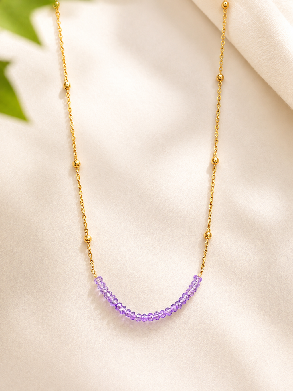 A delicate gold chain necklace featuring a curved row of small, faceted purple beads at the center, displayed on a soft, light beige fabric background.