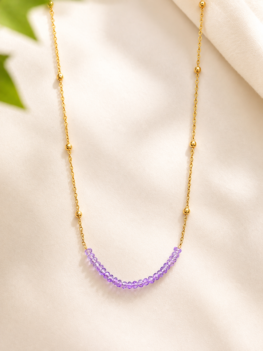 A delicate gold chain necklace featuring a curved row of small, faceted purple beads at the center, displayed on a soft, light beige fabric background.