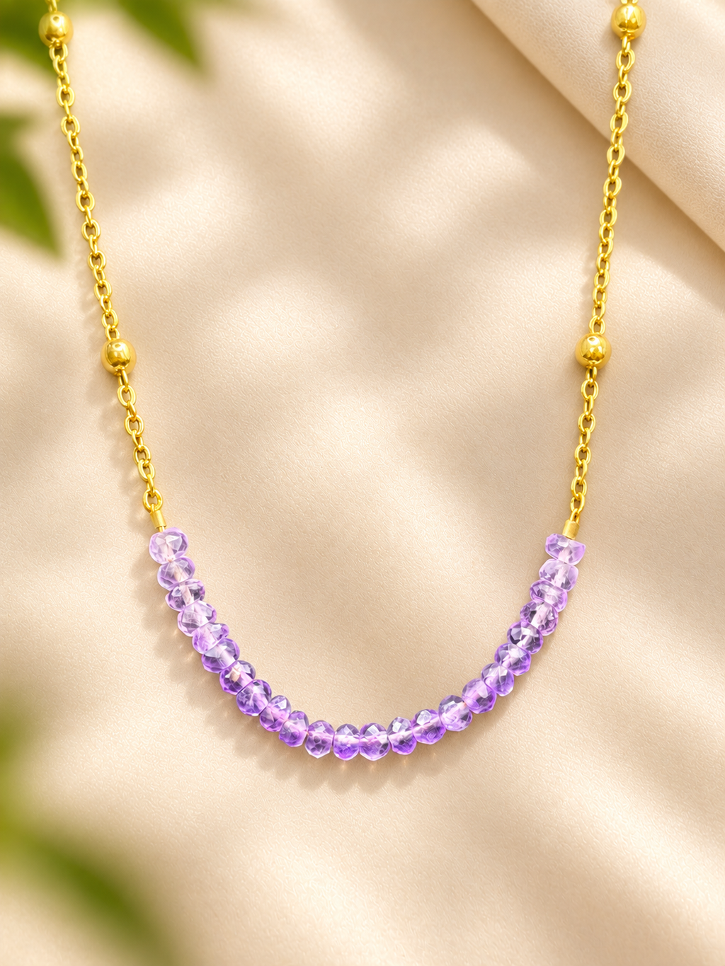 A gold chain necklace featuring a curved row of faceted purple beads, displayed on a soft beige fabric background with subtle green leaf shadows.
