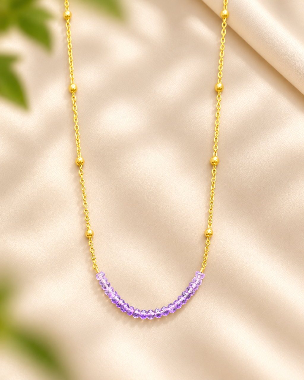 A delicate gold chain necklace with small gold bead accents and a curved row of clear faceted purple beads at the center, displayed on a soft beige satin background with blurred green leaves in the corners.