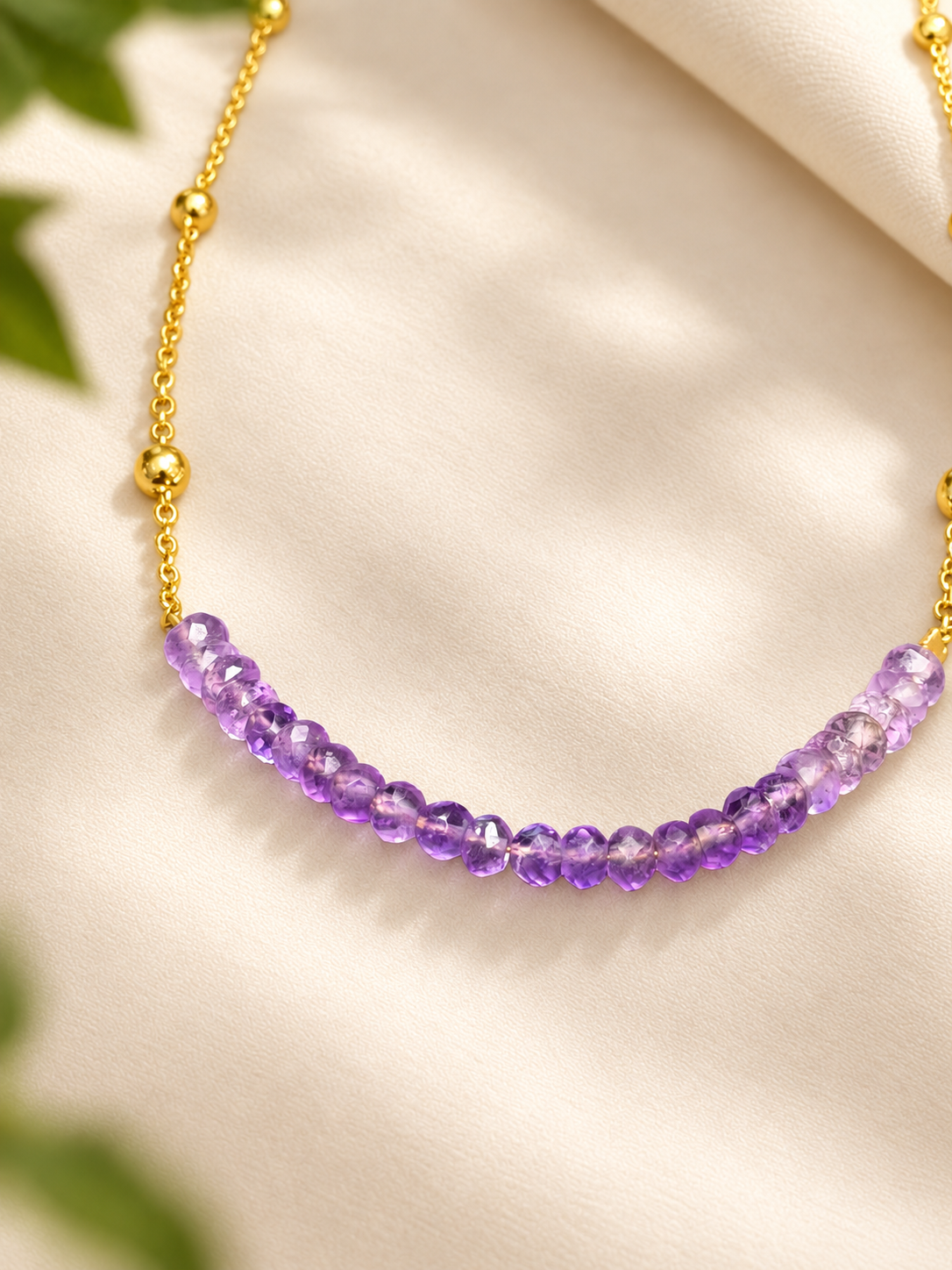 A gold chain necklace featuring a curved row of faceted purple amethyst beads, displayed on a soft cream-colored fabric background with subtle green leaves in the corners.