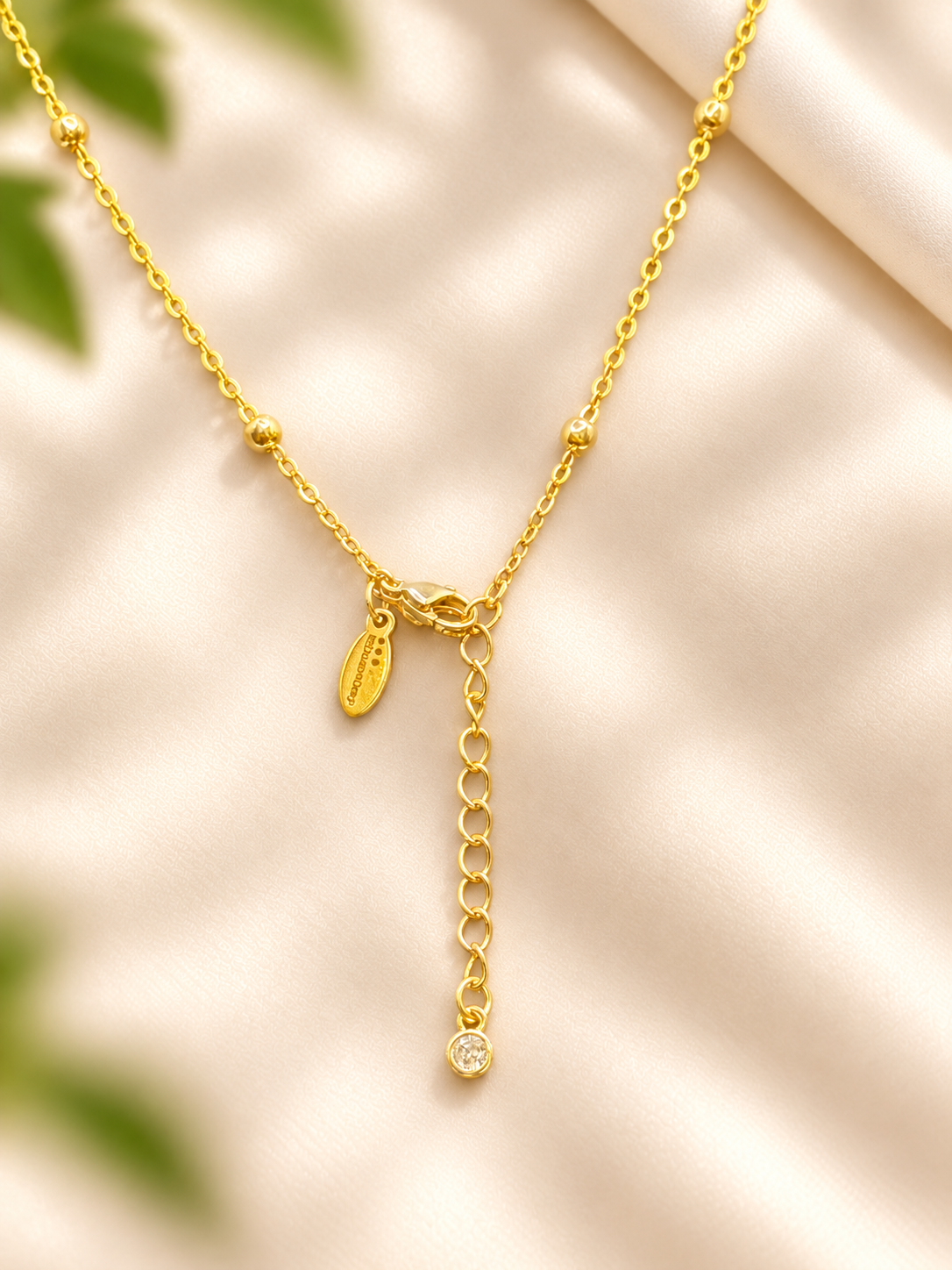 A delicate gold chain necklace with a small oval charm and an adjustable drop extension chain ending in a clear stone, displayed on a soft beige satin fabric background.