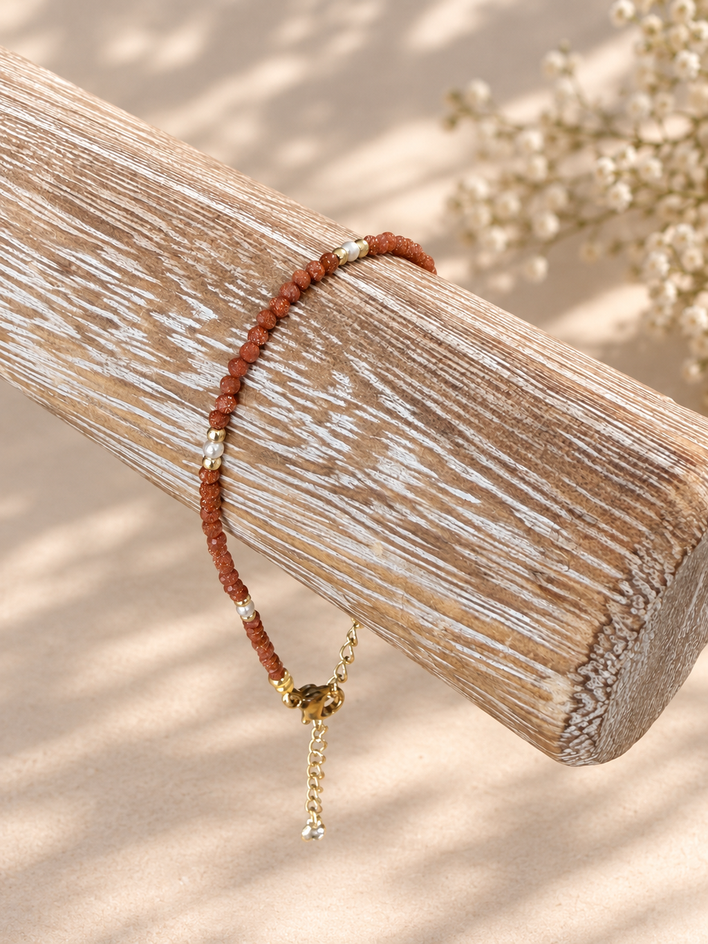 A beaded bracelet with reddish-brown beads, small white pearl-like accents, and a gold-toned charm with a dangling chain, displayed on a weathered wooden log against a warm beige background.