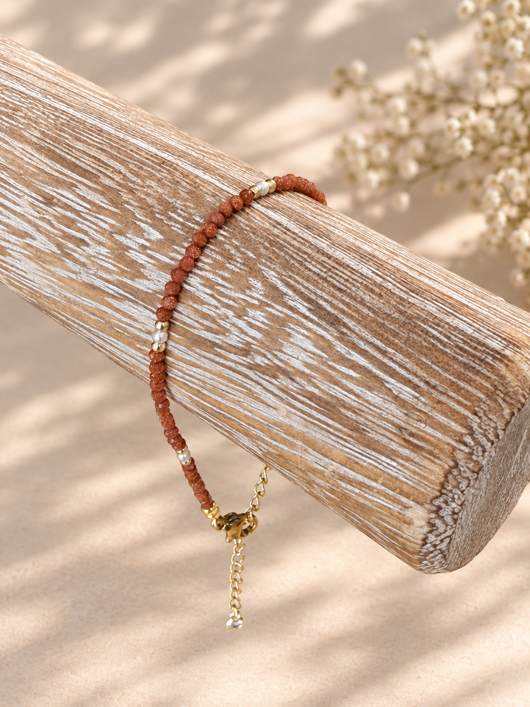 A beaded bracelet with reddish-brown beads, small white pearl-like accents, and a gold-toned charm with a dangling chain, displayed on a weathered wooden log against a warm beige background.