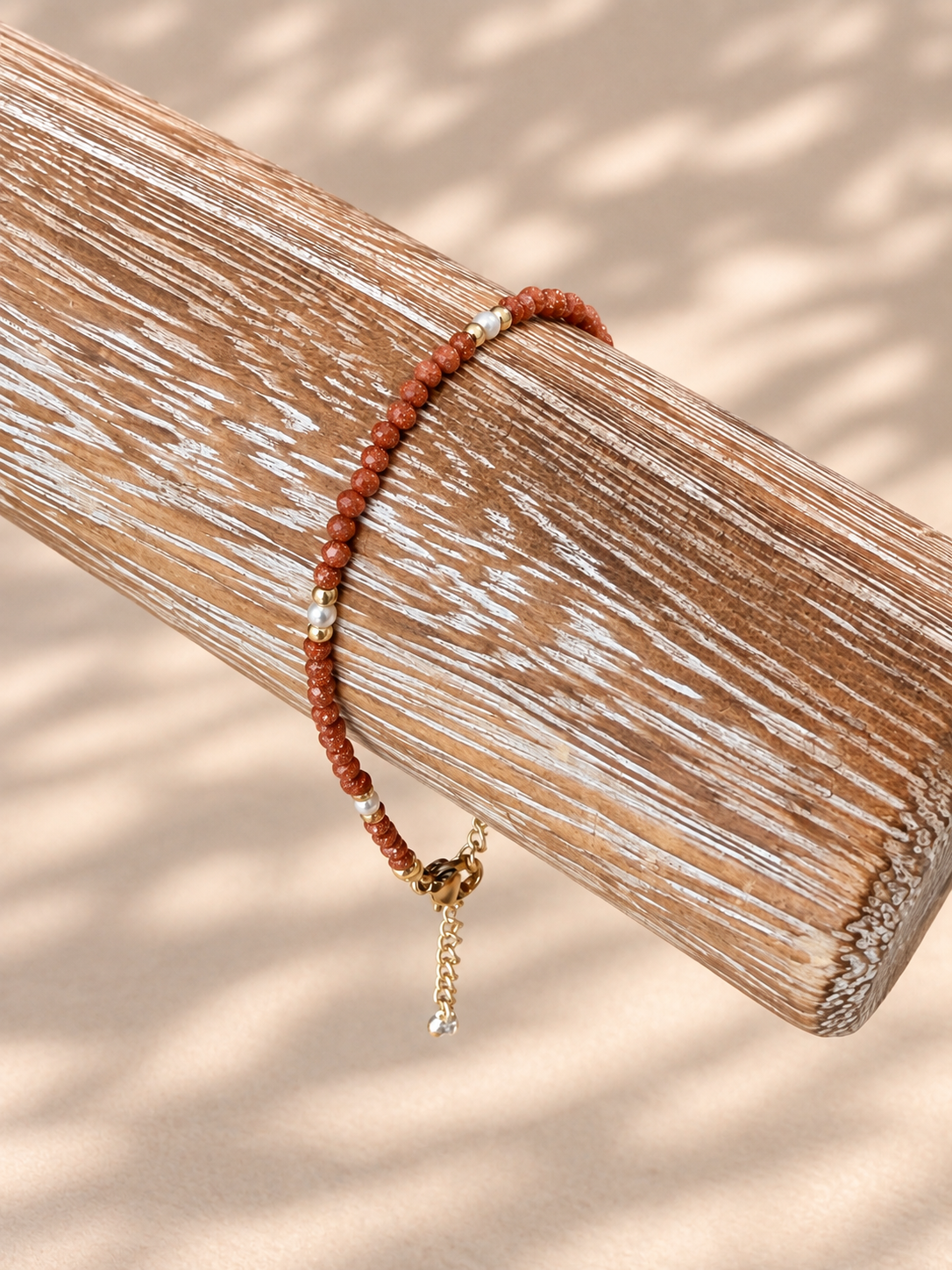A beaded bracelet with terracotta red-orange beads, small white beads, and gold-toned metal accents, displayed on a rustic wooden dowel against a warm beige background with soft leaf shadows.
