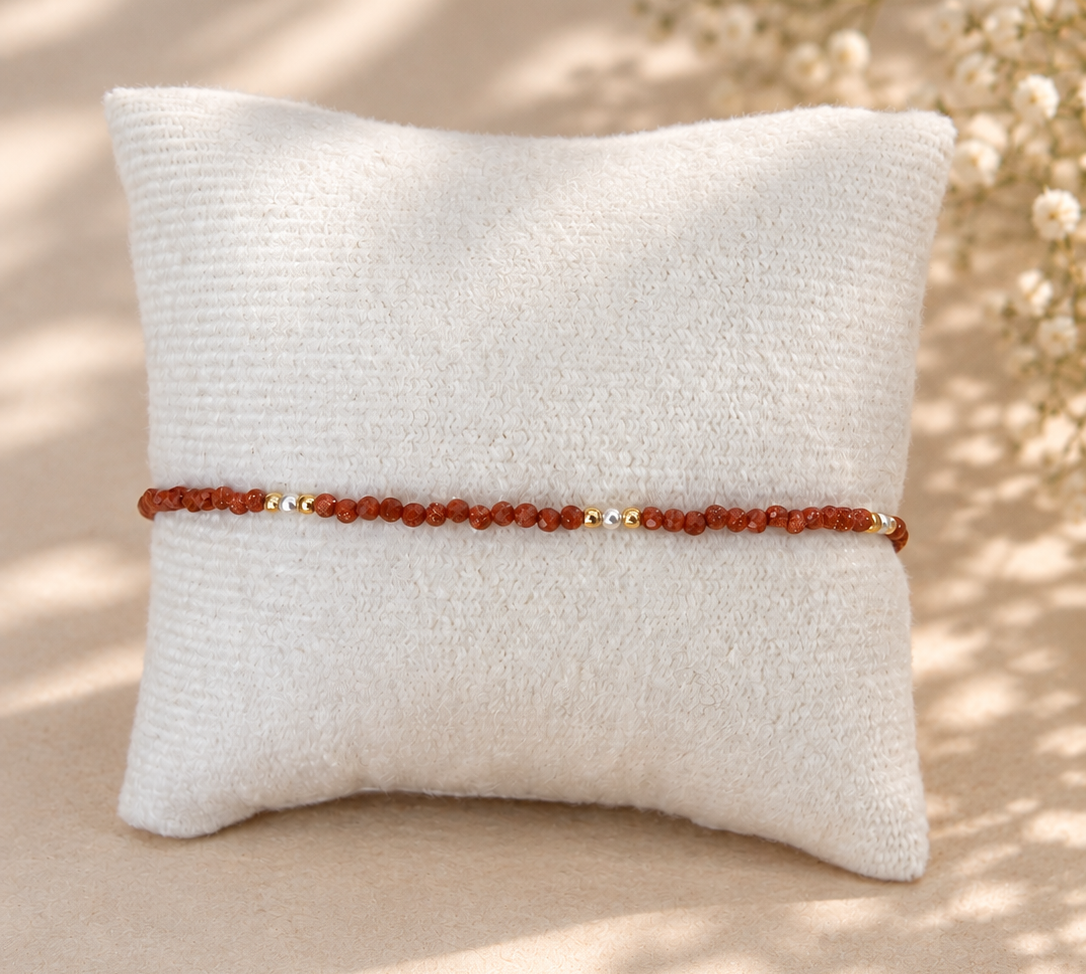 A beaded bracelet with reddish-brown beads and small gold accents, displayed horizontally on a textured off-white pillow against a warm beige background with soft leaf shadows.