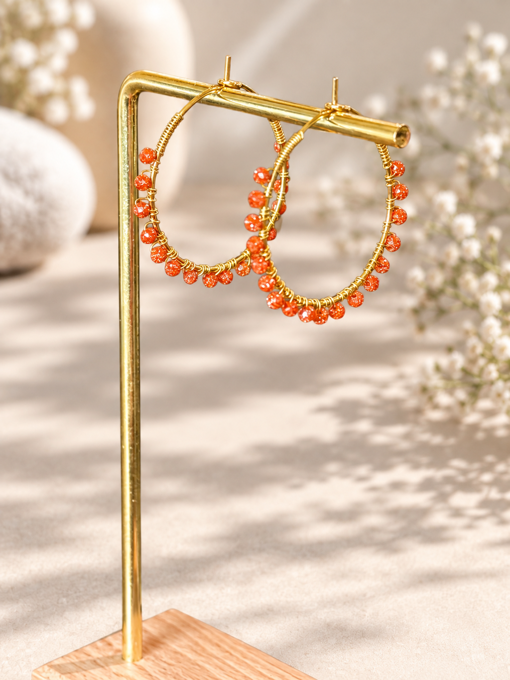Gold hoop earrings with dangling orange bead strands, displayed on a gold stand against a warm beige background with soft floral bokeh.