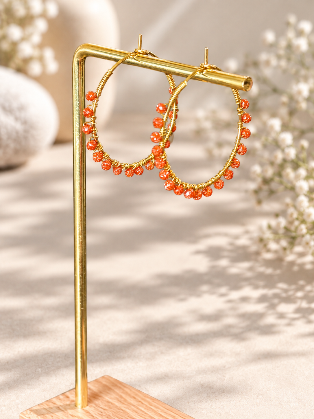 Gold hoop earrings with dangling orange bead strands, displayed on a gold stand against a warm beige background with soft floral bokeh.