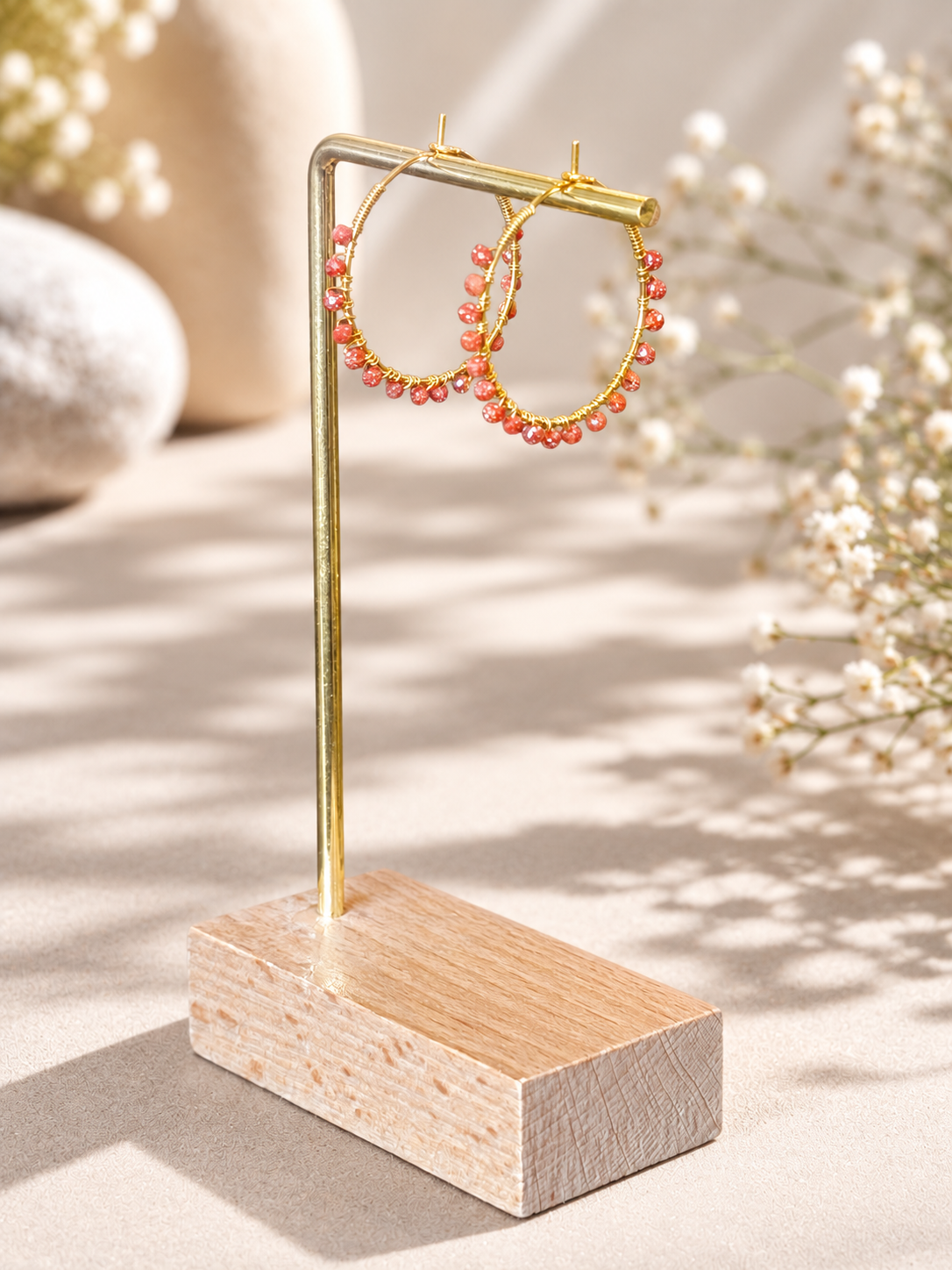 Gold hoop earrings with small coral-pink bead accents, displayed on a gold stand with a wooden base against a warm beige background with soft floral props.