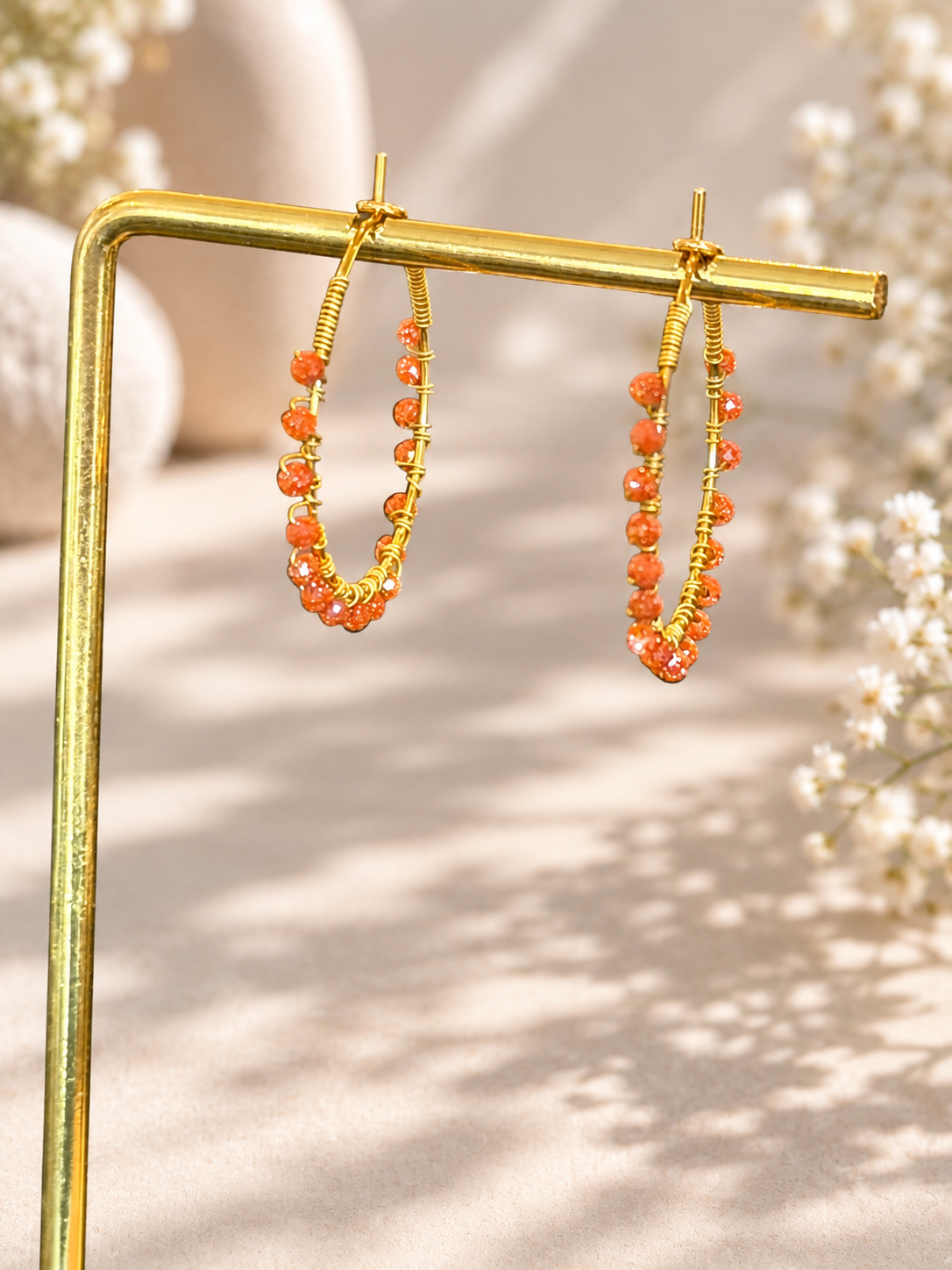 Gold hoop earrings with orange beaded fringe, displayed on a gold stand against a warm beige background with soft floral bokeh.