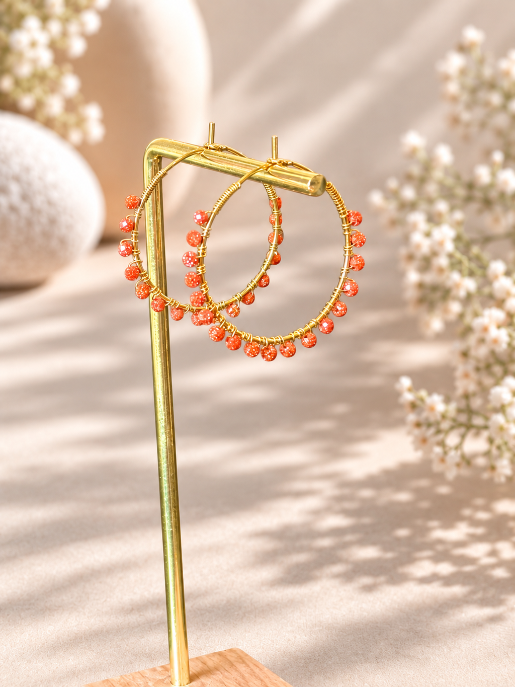 Gold hoop earrings with small orange-red bead strands, displayed on a gold stand against a warm beige background with soft floral bokeh.