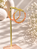 Gold hoop earrings with dangling orange beads, displayed on a gold stand with a wooden base against a softly blurred beige background with white flowers.