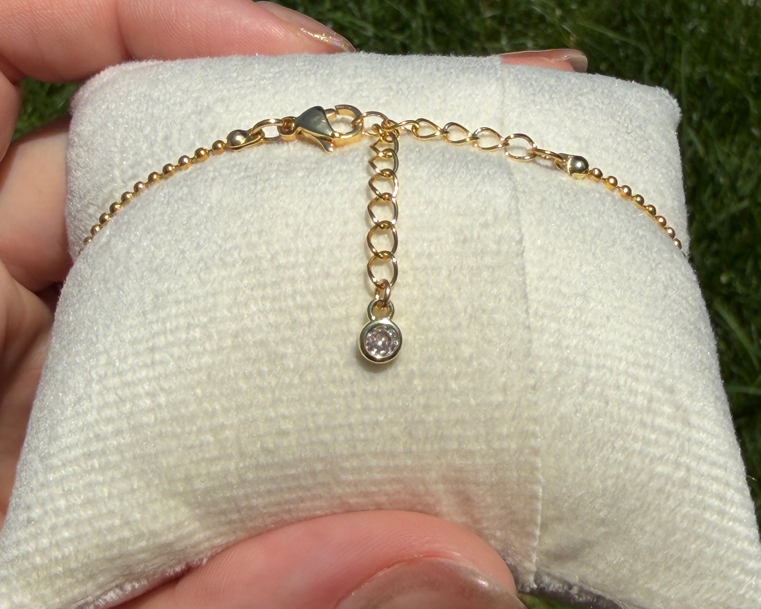 Gold bracelet with a small gemstone on a white cushion, held by a hand against a grassy background.