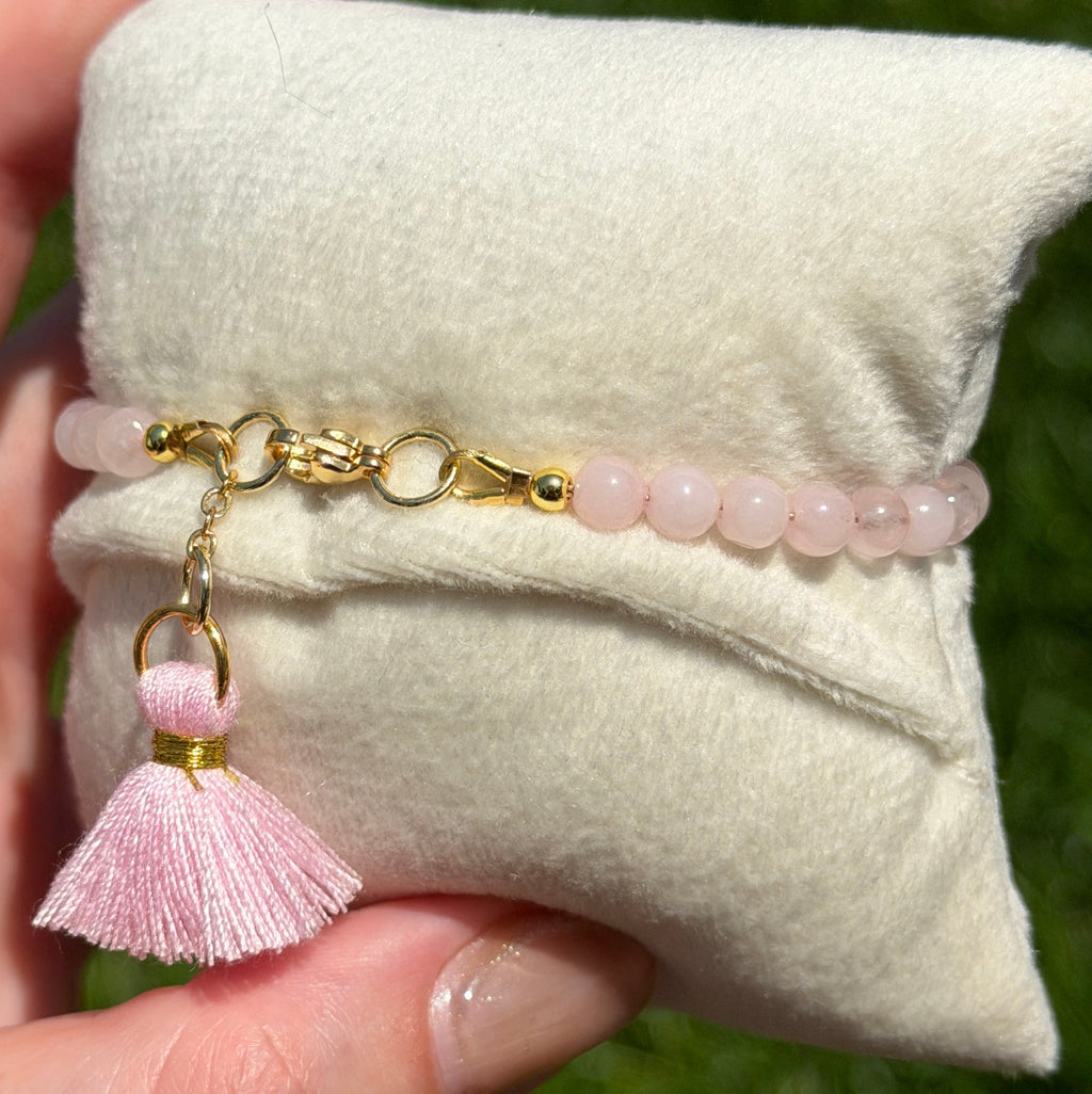 Hand holding a small beige pillow with a pink bracelet featuring beads and tassel against a grassy background