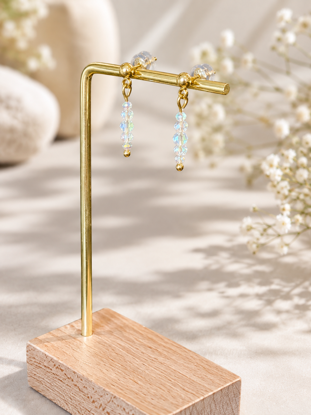 A pair of gold-tone dangle earrings with iridescent blue and white beads, displayed on a gold stand with a wooden base against a softly blurred neutral background with white flowers.