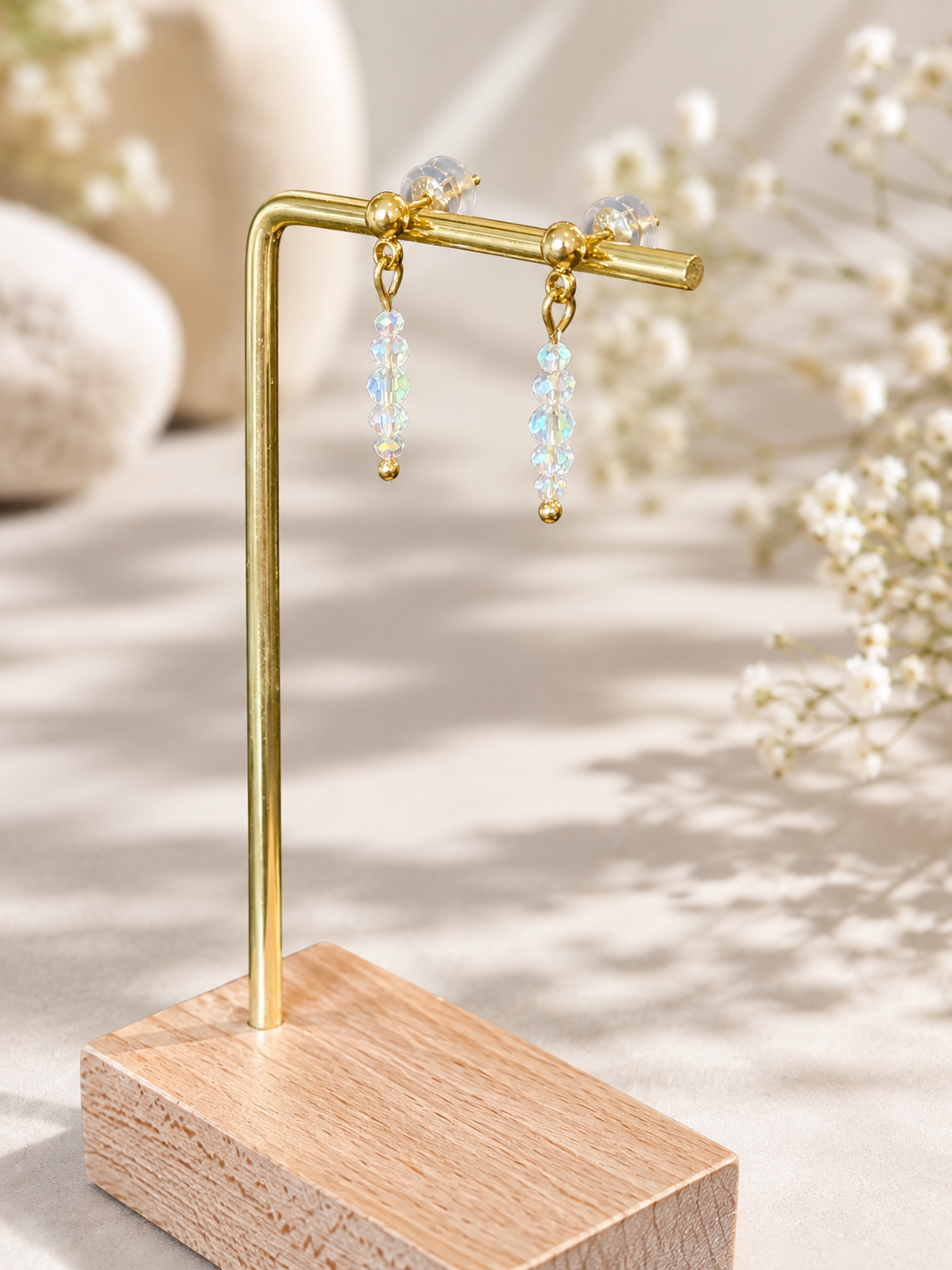 A pair of gold-tone dangle earrings with iridescent blue and white beads, displayed on a gold stand with a wooden base against a softly blurred neutral background with white flowers.