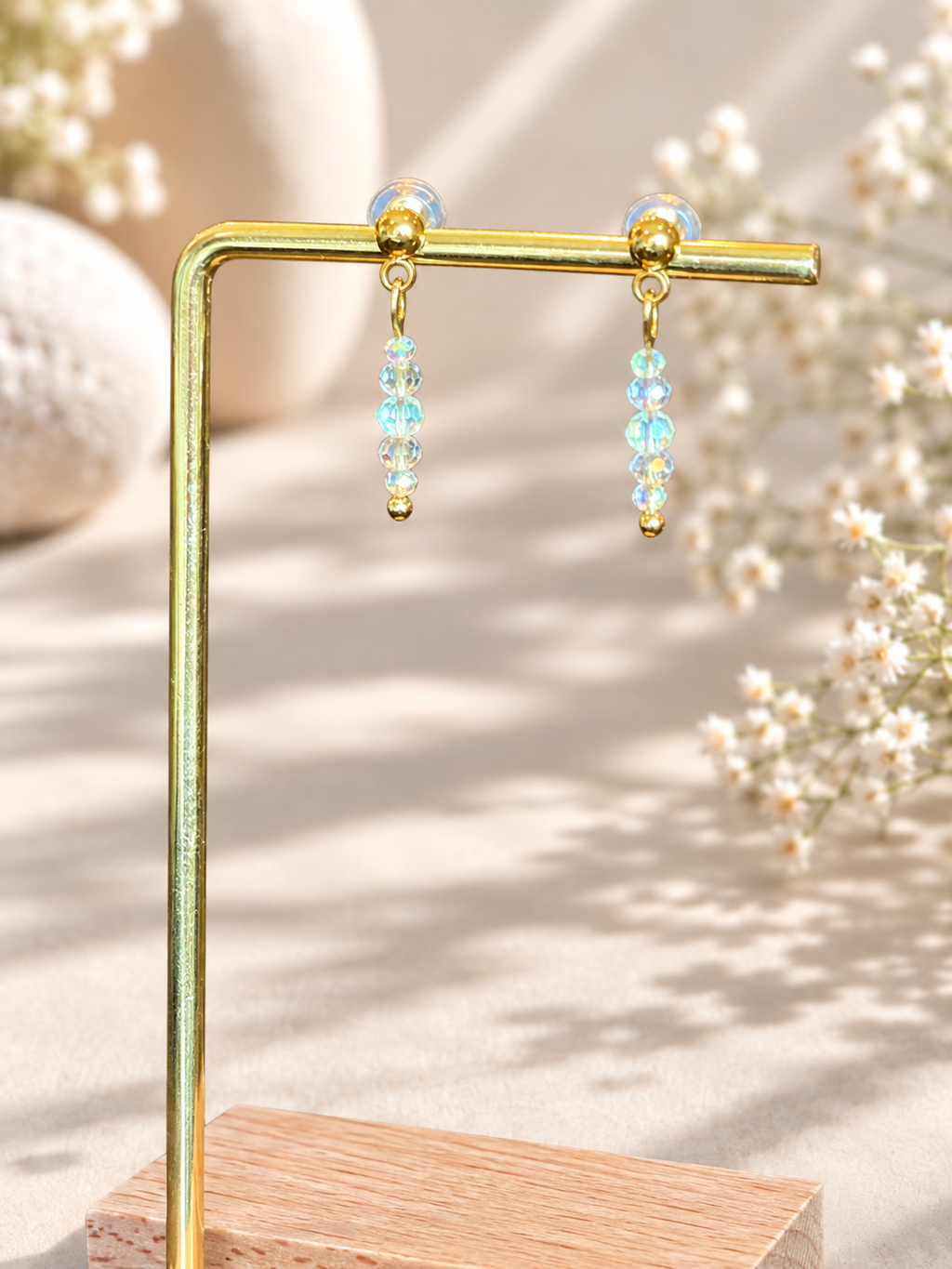 A pair of gold-tone dangle earrings with iridescent blue-green beads, displayed on a gold metal stand against a warm beige background with soft floral props.