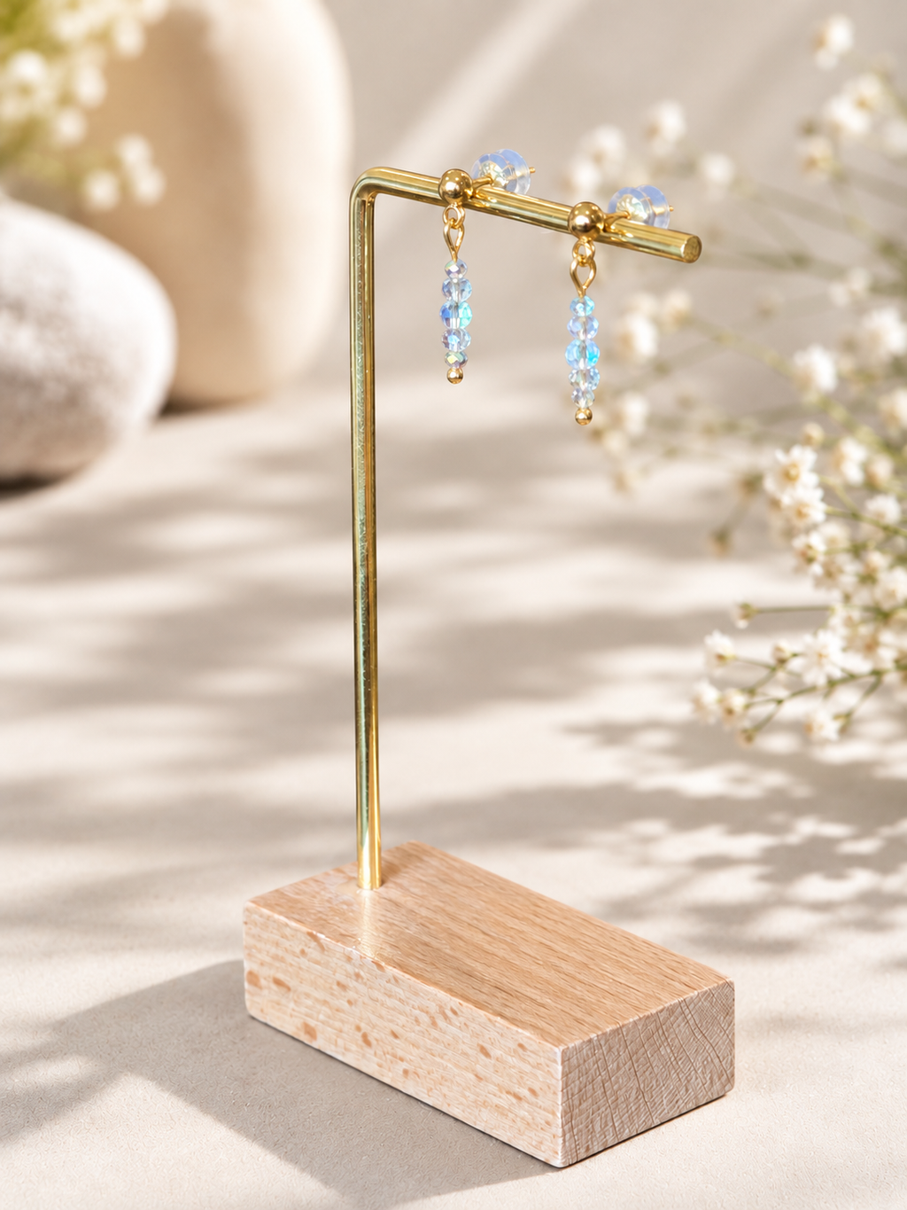 Gold-tone dangle earrings with light blue faceted beads displayed on a wooden stand with a brass S-shaped hook, set against a warm beige background with soft floral bokeh.