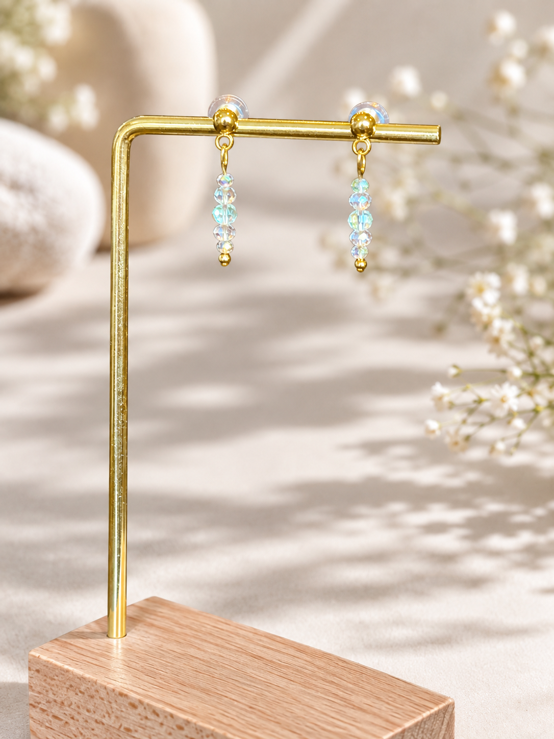 A pair of gold drop earrings with light blue and iridescent beads, displayed on a gold L-shaped stand over a wooden base against a softly blurred neutral background with white flowers.