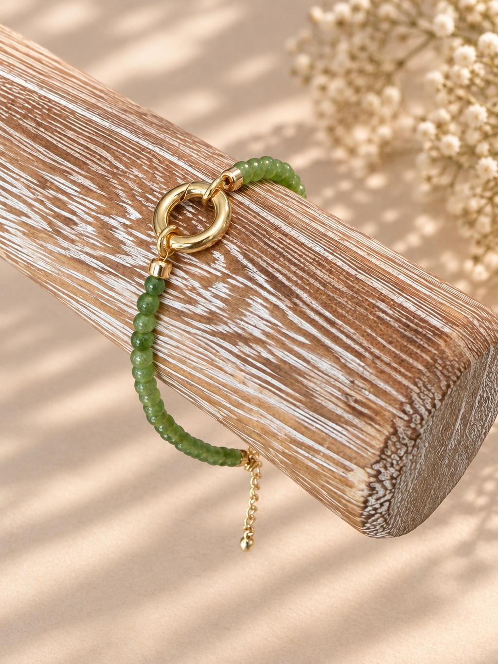 A green beaded bracelet with a gold ring clasp and small gold chain detail, displayed on a textured wooden block with soft beige shadows and dried flowers in the background.
