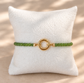 A green beaded bracelet with a gold circular ring clasp, displayed on a white textured pillow against a beige background with soft dappled shadows.
