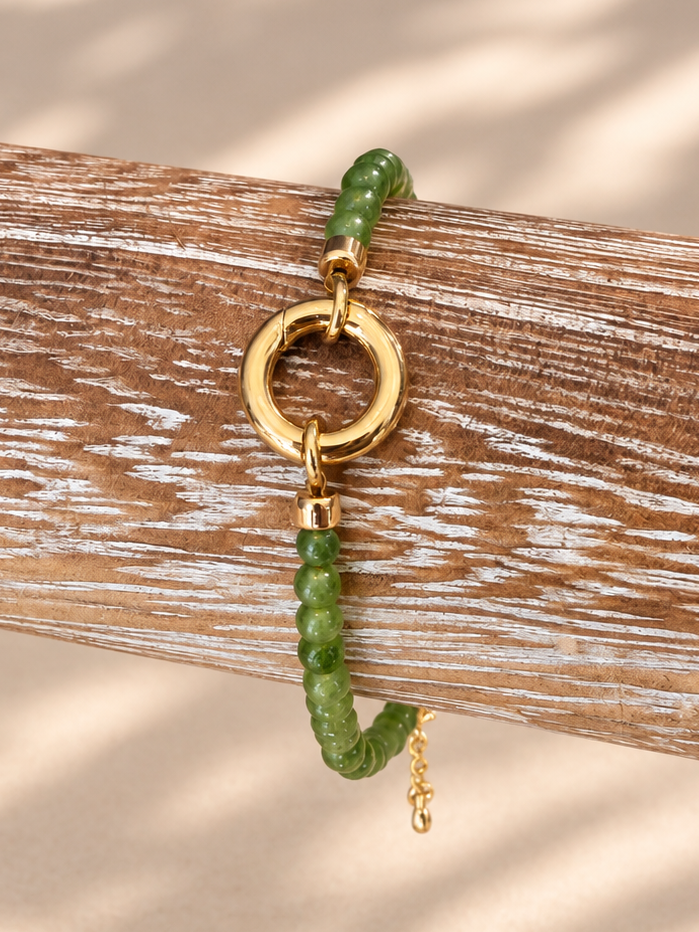 A beaded bracelet with green beads, a gold ring clasp, and a dangling green bead charm, displayed on a weathered wooden branch against a beige background with soft leaf shadows.
