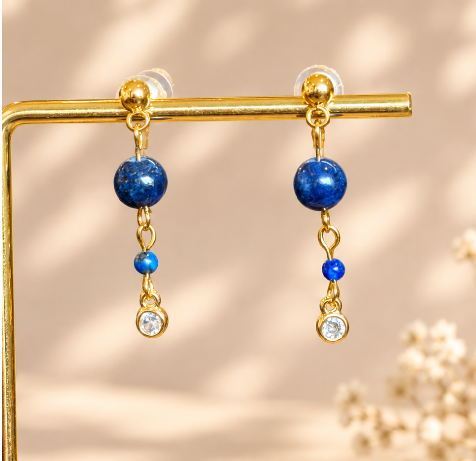 Gold dangle earrings with a large blue bead, a small blue bead, and a clear crystal charm, displayed on a gold stand against a warm beige background with soft shadows.