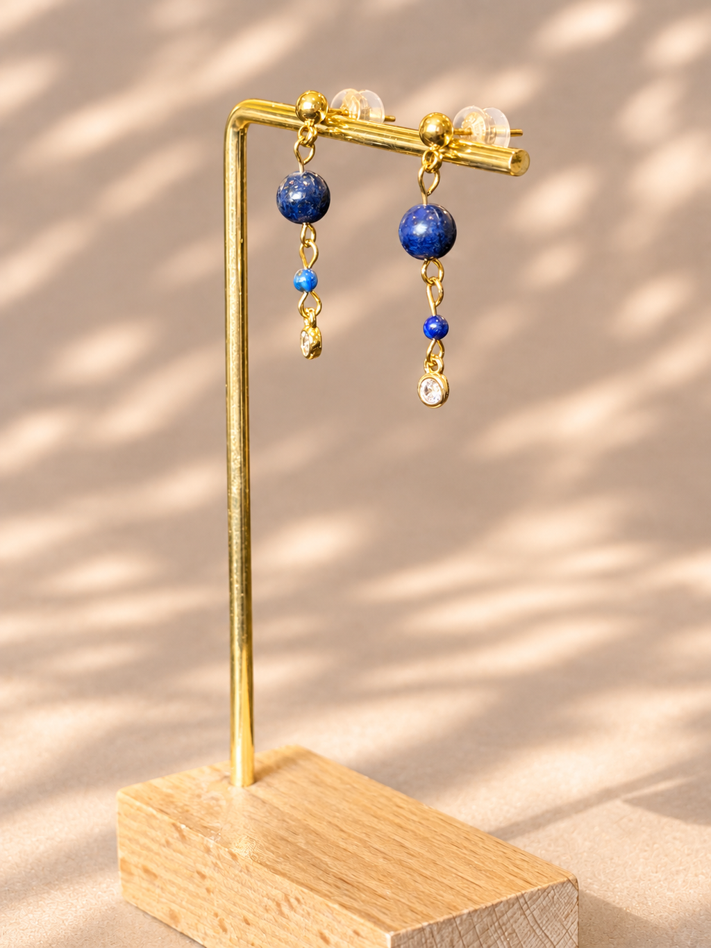 Gold-tone dangle earrings with blue beads and clear crystal drops, displayed on a gold stand over a wooden base against a beige background with soft shadow patterns.