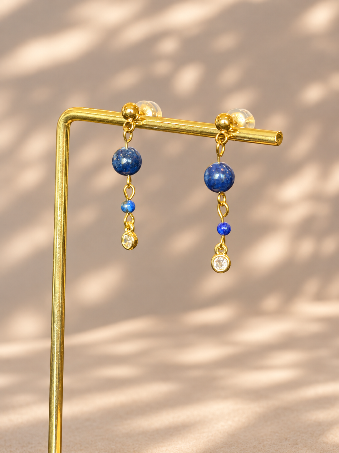 A pair of gold dangle earrings with blue beads and clear gemstones, hanging from a gold display stand against a beige background with soft leaf shadows.