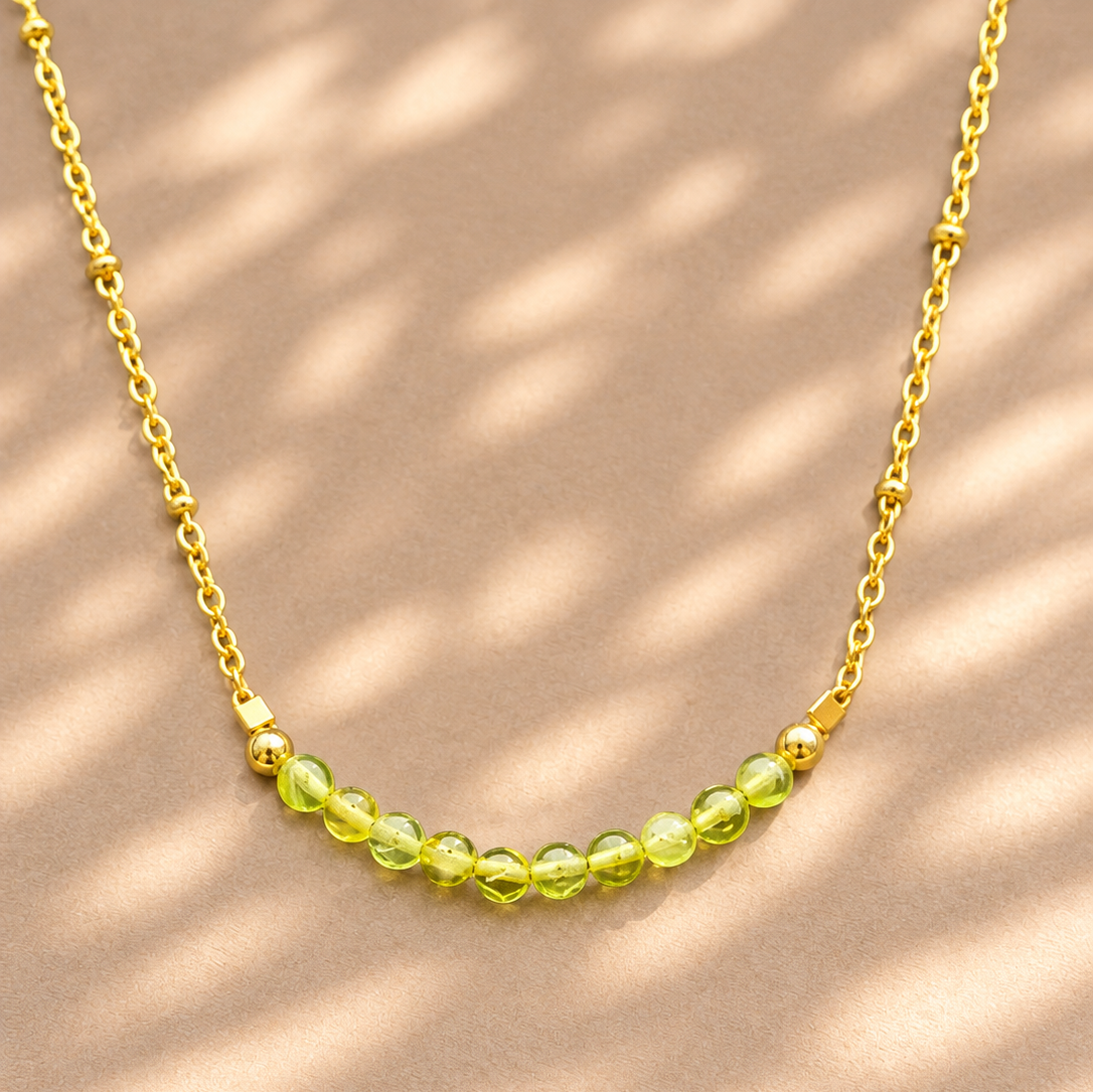 A delicate gold chain necklace featuring a curved row of small, faceted lime-green beads, displayed on a warm beige surface with soft, dappled shadows.