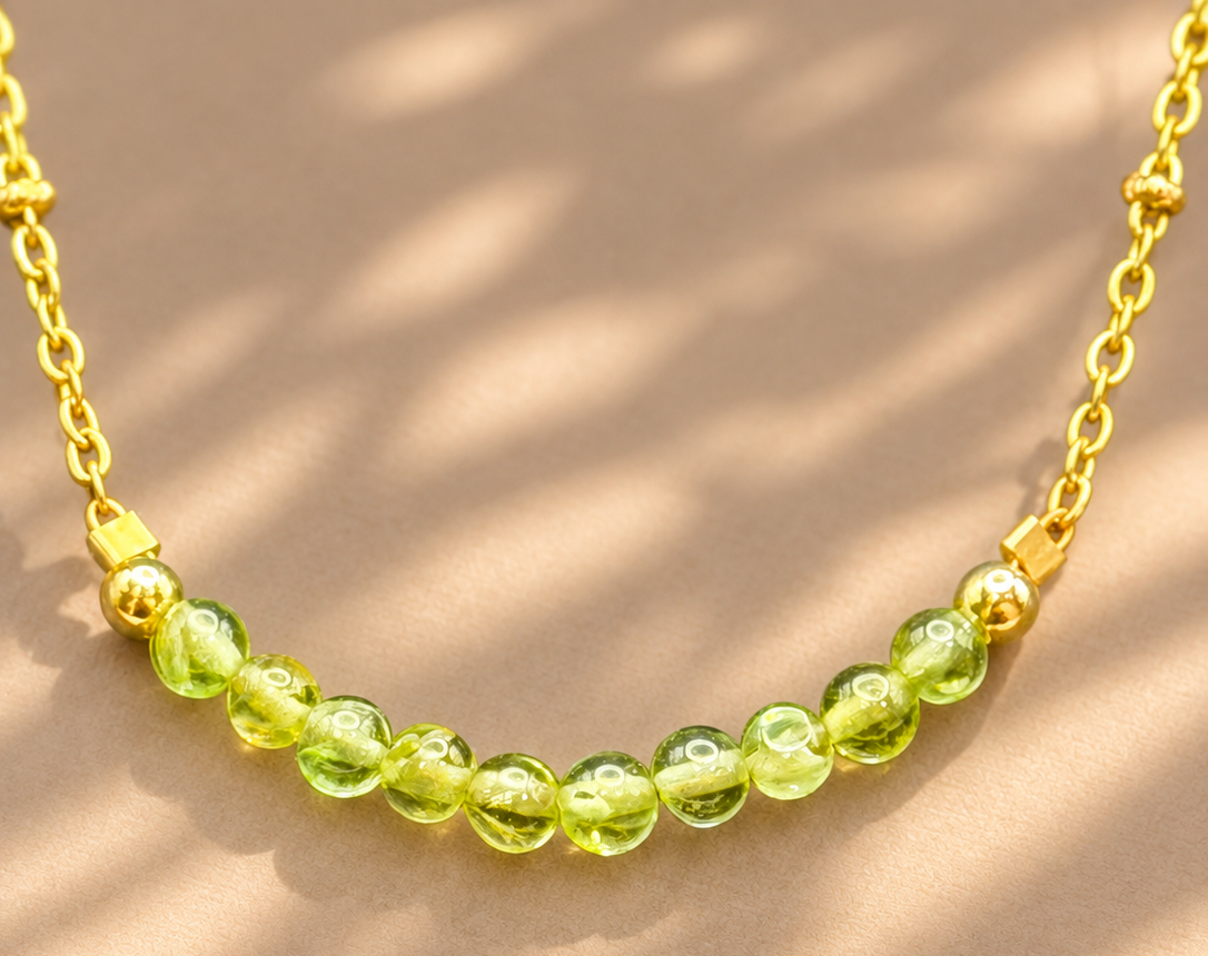 A gold chain necklace featuring a curved row of translucent light green round beads, displayed on a warm beige surface with soft dappled shadows.