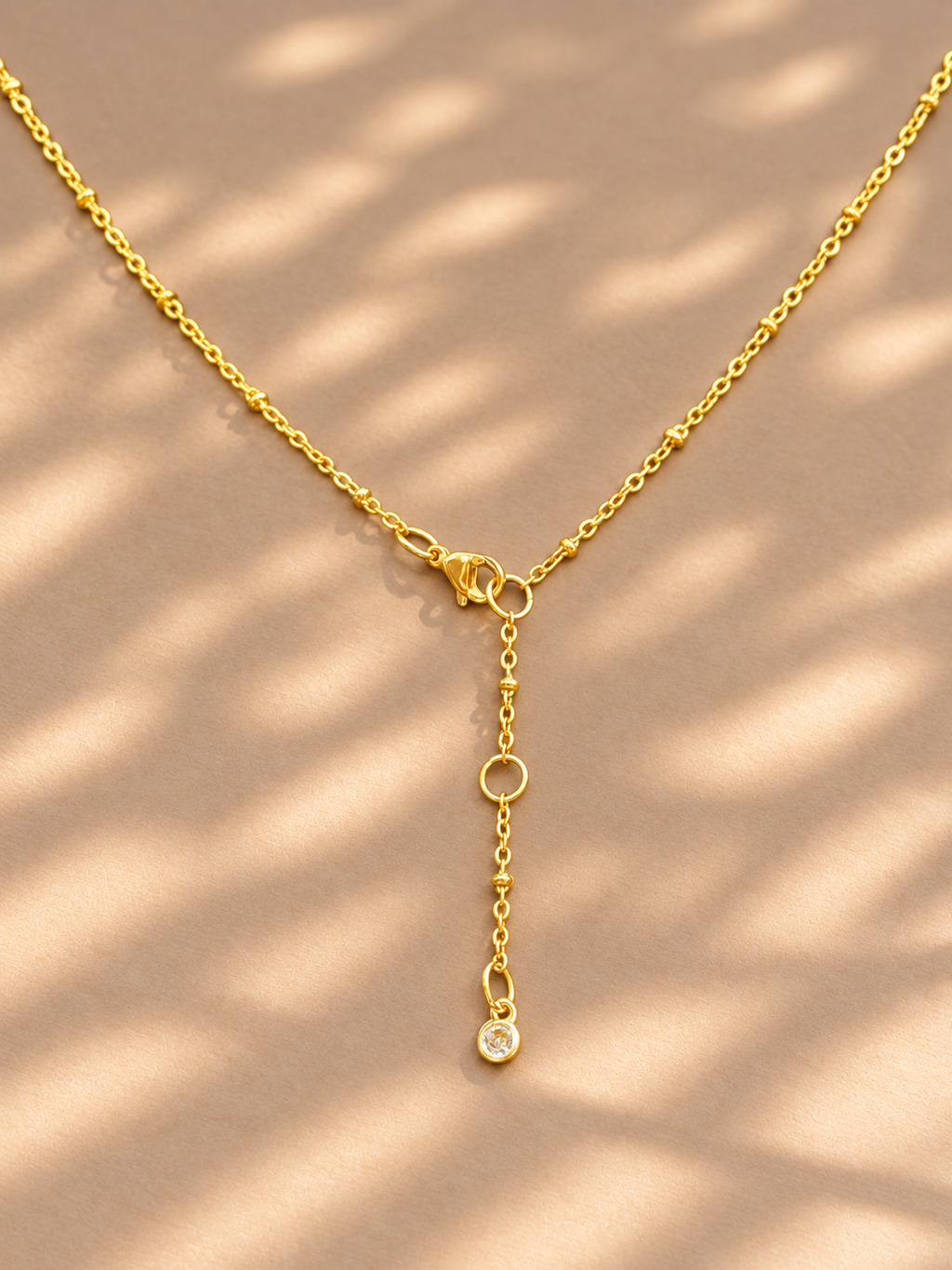 A delicate gold Y-shaped necklace with a small clear gemstone pendant, displayed on a warm beige surface with soft leaf shadow patterns.