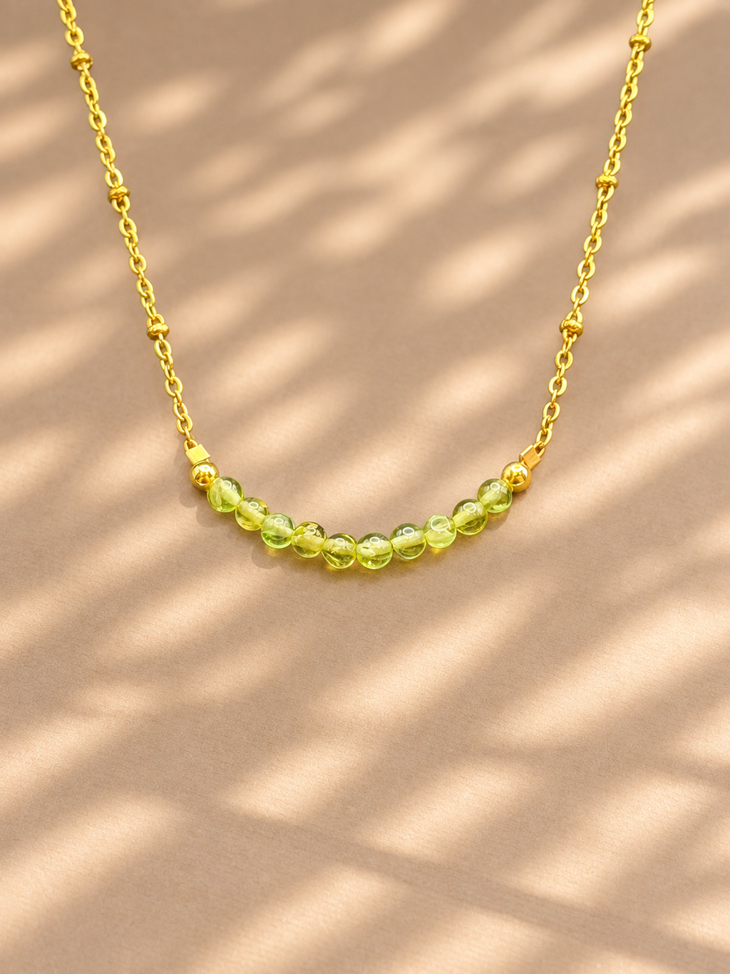 A delicate gold chain necklace featuring a curved row of faceted light green beads, displayed on a warm beige surface with soft shadow overlays.