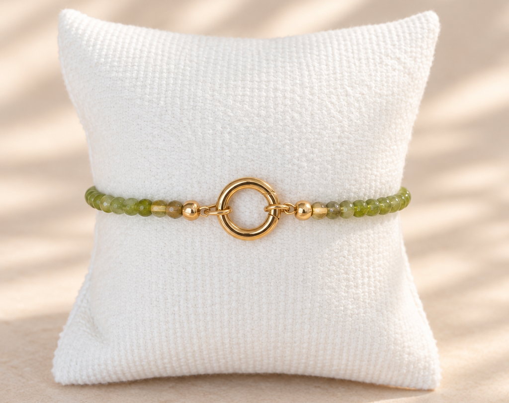 A beaded bracelet with light green and gold beads, featuring a central gold ring clasp, displayed on a white textured pillow against a warm beige background with soft leaf shadows.