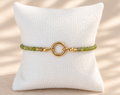 A beaded bracelet with light green and gold beads, featuring a central gold ring clasp, displayed on a white textured pillow against a warm beige background with soft leaf shadows.