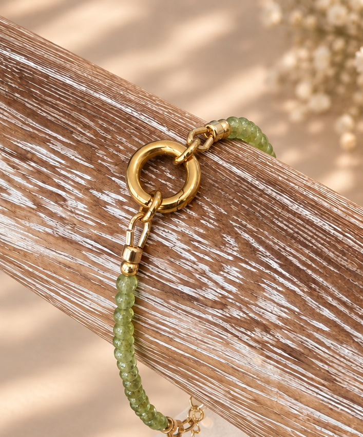 A green beaded bracelet with a gold ring clasp and charm, displayed on a rustic wooden stand with a small paper tag.