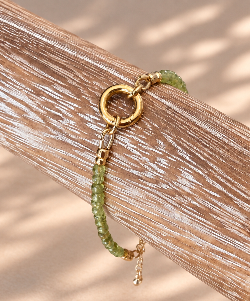 A gold-tone beaded bracelet with a circular ring clasp and a small dangling charm, draped over a rustic wooden branch on a beige surface with soft leaf shadows.