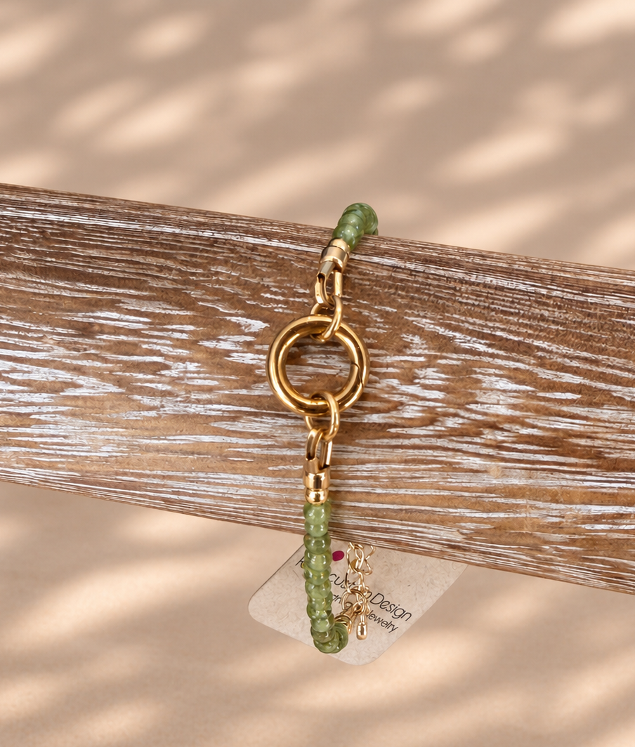 A gold-tone keychain charm with green beads and a small hanging tag, displayed on a weathered wooden block against a beige background with soft leaf shadows.