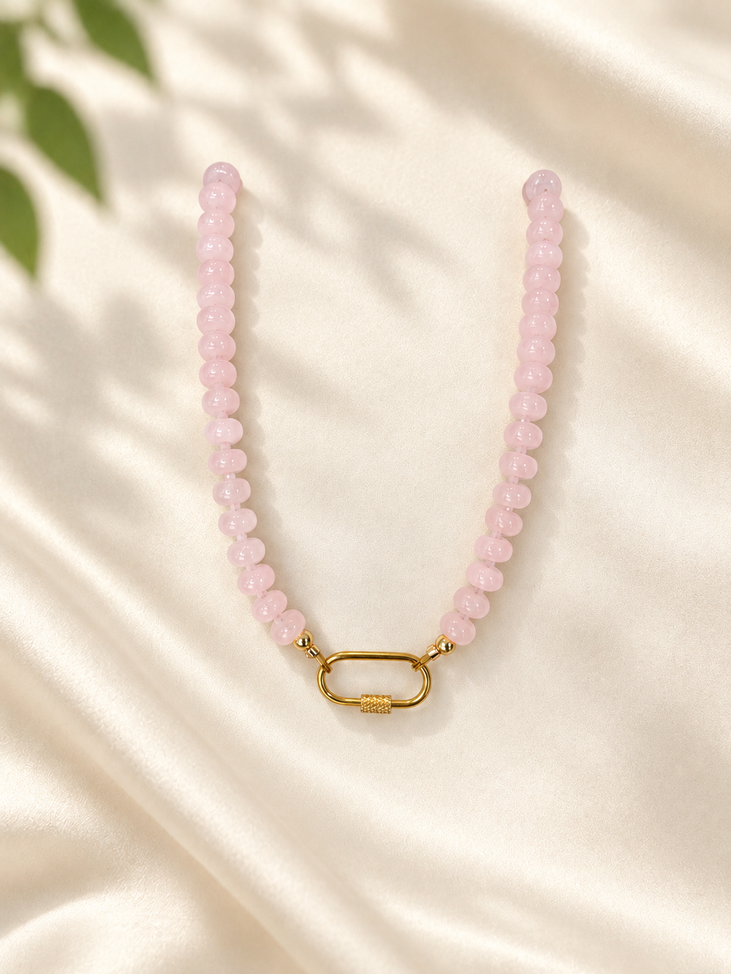 A pink beaded necklace with a gold clasp, displayed on a cream satin fabric background with soft leaf shadows.