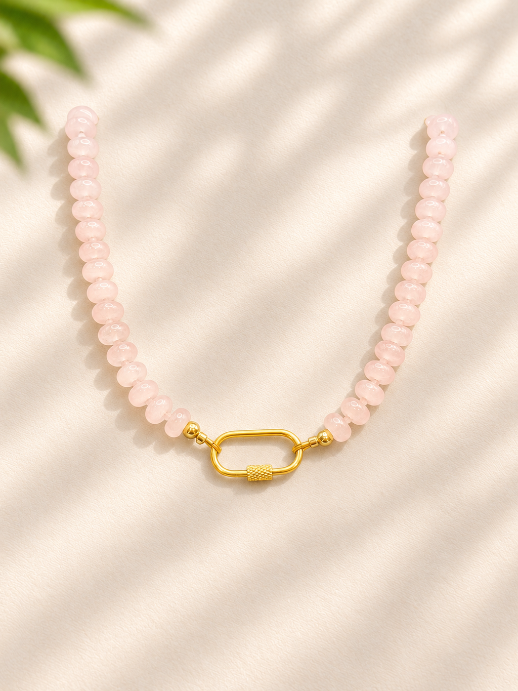 A beaded necklace made of light pink faceted beads with a textured gold clasp, displayed on a cream-colored fabric background with soft leaf shadows.